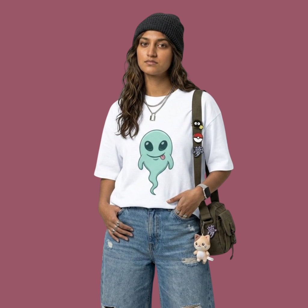 Front view of a female model wearing an oversized white t-shirt featuring a cute green alien ghost graphic from the anime Dan Da Dan. She is styled with blue jeans, a dark grey beanie, and a crossbody bag decorated with anime pins and a small cat plushie.