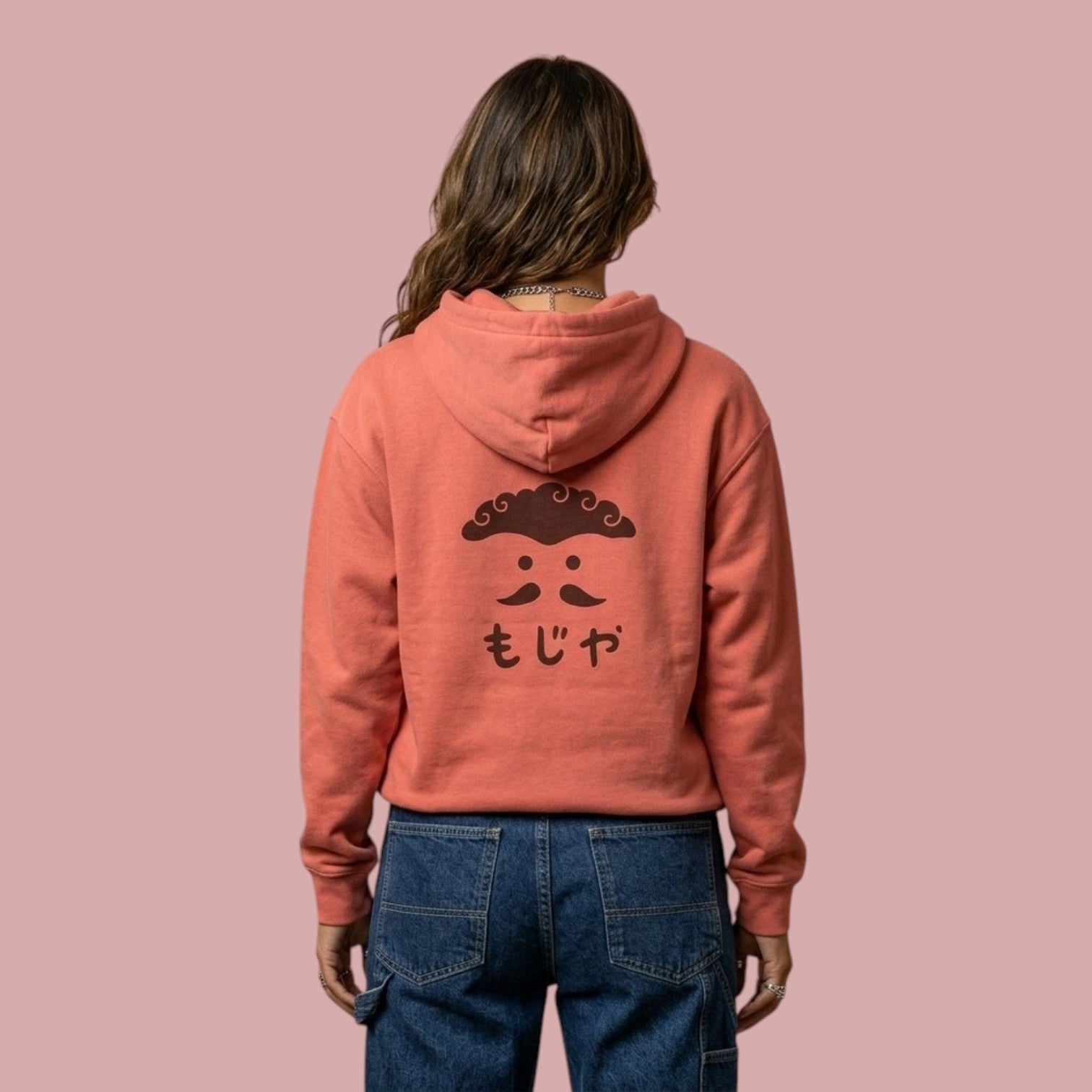 Back view of a female model wearing a peach-colored Dan Da Dan anime hoodie. The back features a black graphic design of a stylized face with curly hair, dotted eyes, a mustache, and Japanese text reading "Moja" underneath