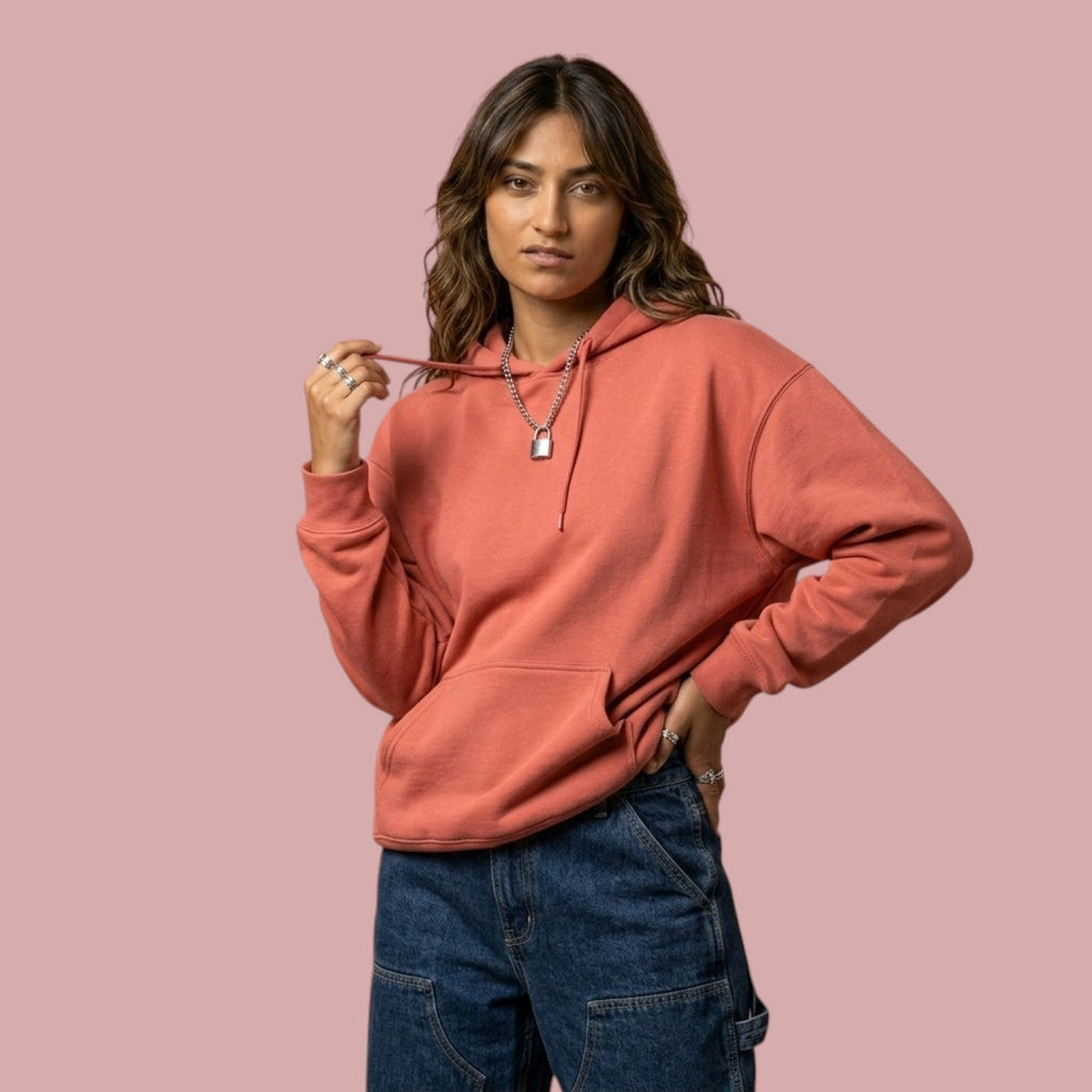 A female model facing forward wearing a plain, peach-colored pullover streetwear hoodie inspired by the Dan Da Dan anime. She is styling the hoodie with blue jeans and silver jewelry against a pink background.