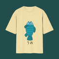 Front view of a pale yellow t-shirt from the Dandadan collection, featuring Momo Ayase's iconic blue cartoon fish design and Japanese text