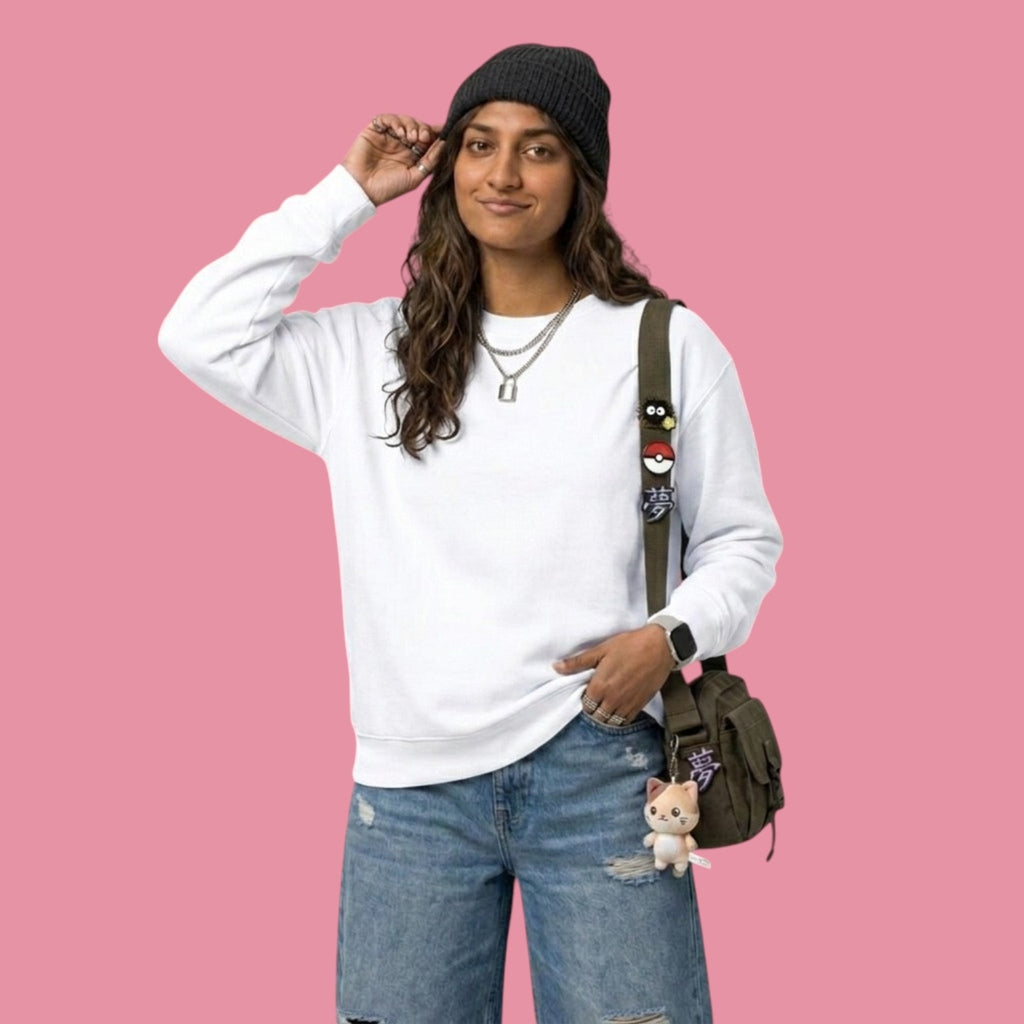Front view of a model smiling while wearing a classic white crewneck sweatshirt from the Dan da Dan streetwear collection. The model is wearing a black beanie, silver necklaces, baggy blue jeans, and a green messenger bag decorated with anime pins and a cat plushie