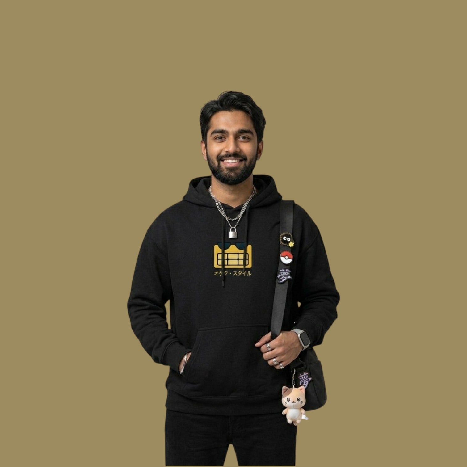 Smiling male model wearing a black Dan Da Dan inspired hoodie featuring a yellow pixelated alien graphic and the Japanese text "Otaku Style". He is accessorized with a silver padlock necklace and a black crossbody bag decorated with anime pins and a cat plush keychain.