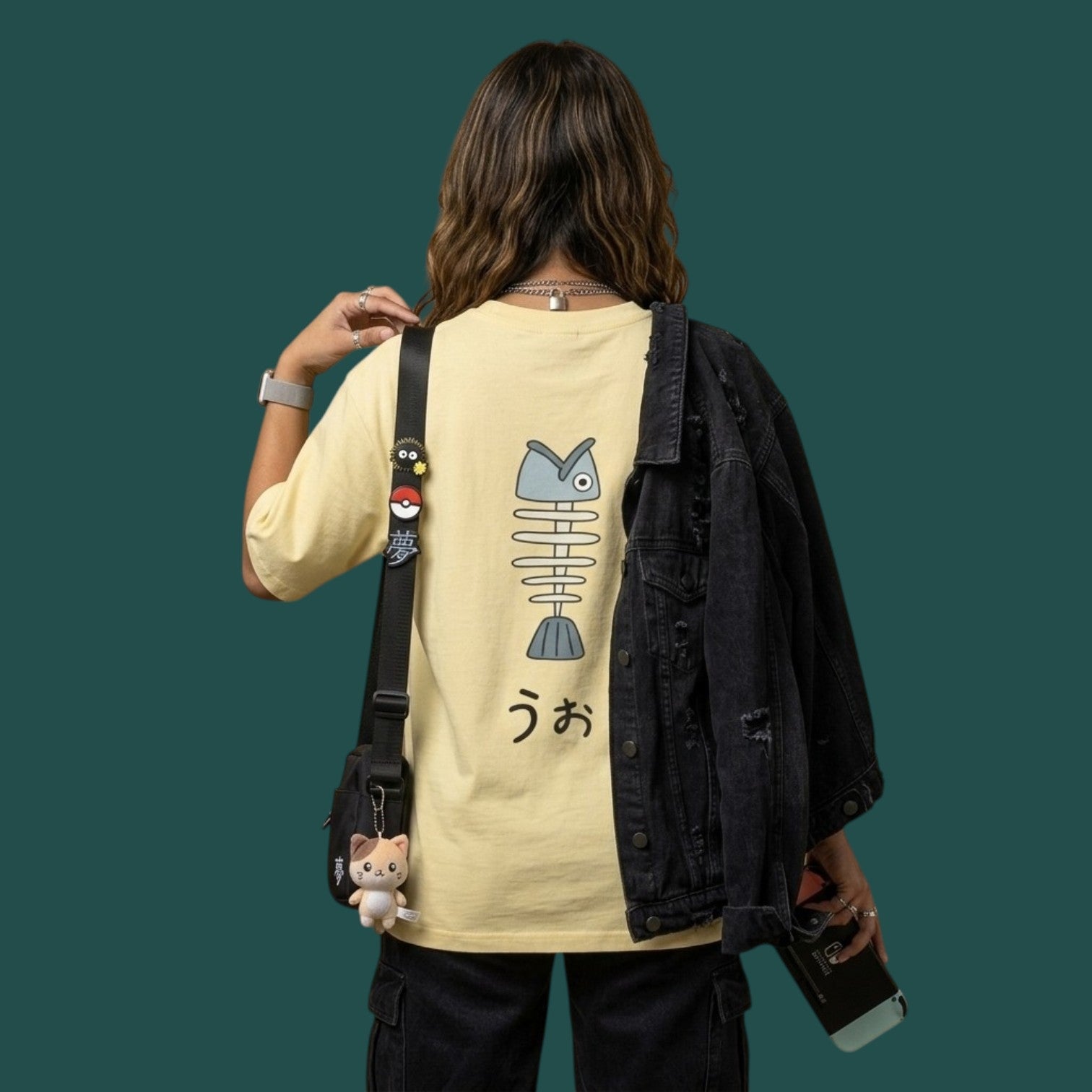 Back view of a model wearing the yellow Dandadan streetwear t-shirt, showcasing the fish skeleton graphic on the back. She has a black denim jacket slung over one shoulder and is wearing a crossbody bag with anime keychains.