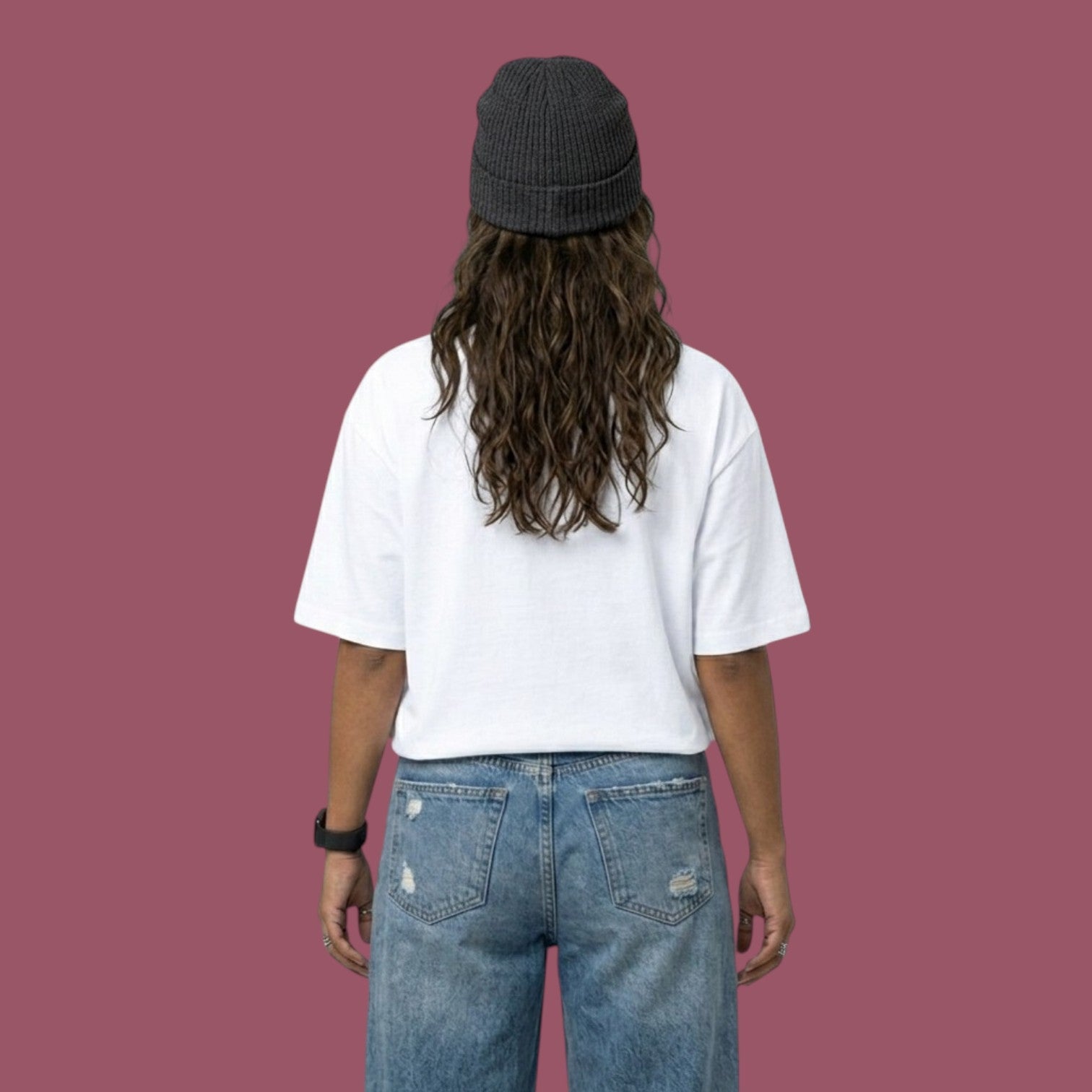 Back view of a female model wearing an oversized plain white t-shirt, baggy distressed blue jeans, and a dark grey beanie against a solid mauve background.