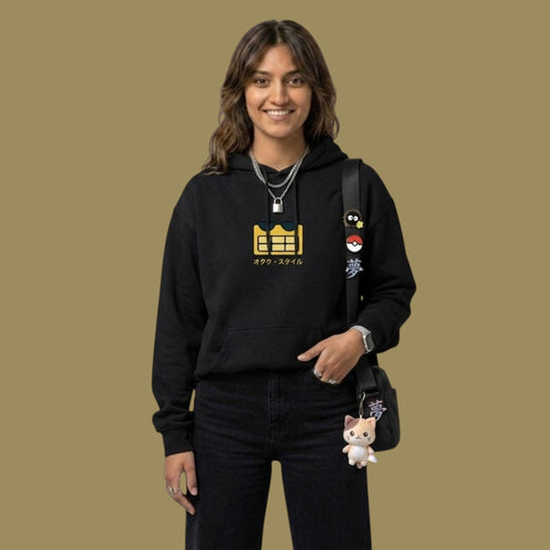 Smiling female model looking at the camera wearing a black Dan Da Dan streetwear hoodie with a yellow pixelated alien logo and Japanese text. She is carrying a black crossbody bag accessorized with anime pins and a small cat plushie.