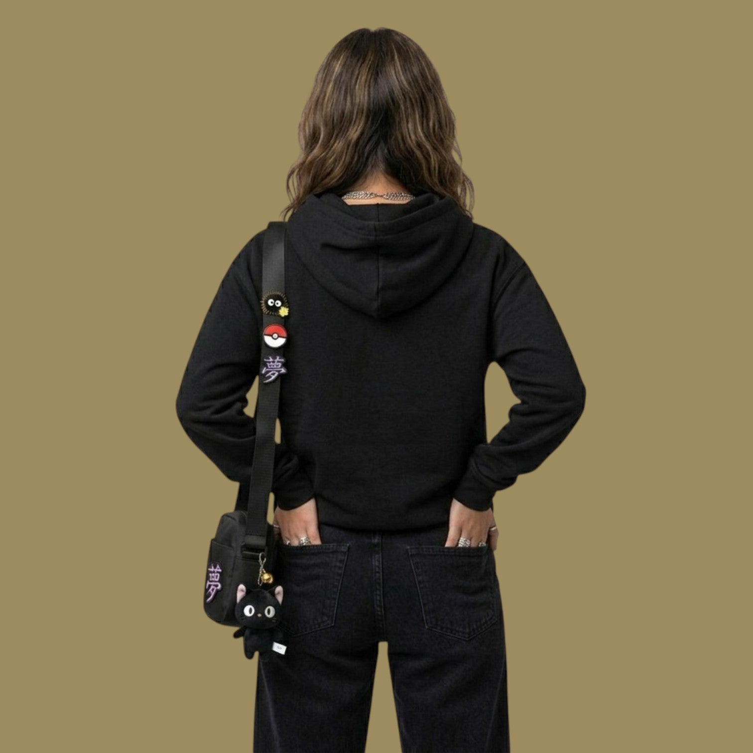 Back view of a female model styling a black Dan Da Dan inspired hoodie with black denim. She has her hands in her pockets and is wearing a black crossbody strap adorned with anime pins and a black cat plush keychain.